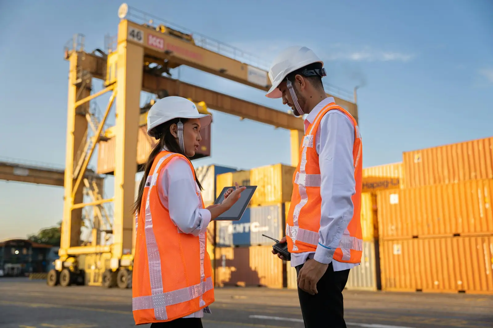 Two port workers in hi-vis reviewing a plan on a tablet beside stacked containers.
